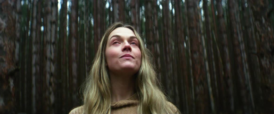 A young woman stands in a forest full of tall trees while looking skyward