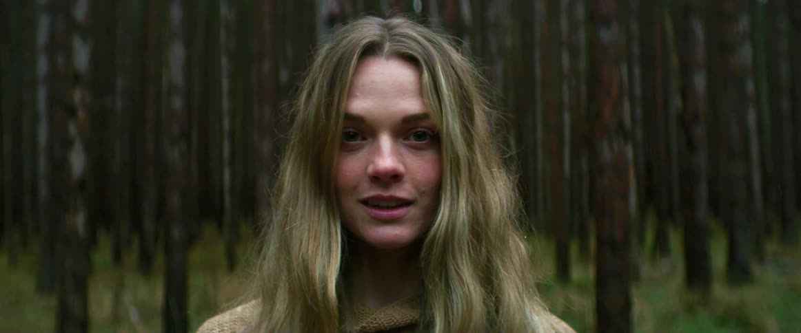 A woman smirks and smiles as she stands in the woods