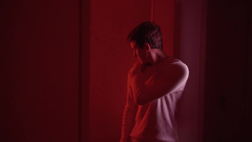 A young man stands indoors looking pensive as a red light subtle strobes on and off nearby