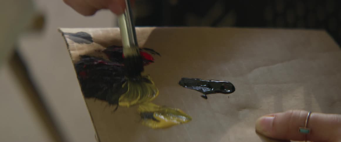 A person in a room mixing paint on a piece of cardboard with a paint brush with a canvas and wall in the background