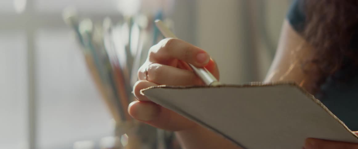 A woman holds a paint brush and palette blending color near a window in an art studio