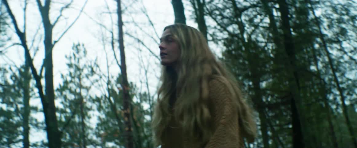 A woman with a sweater walking in the forest with trees the sky and clouds in the background