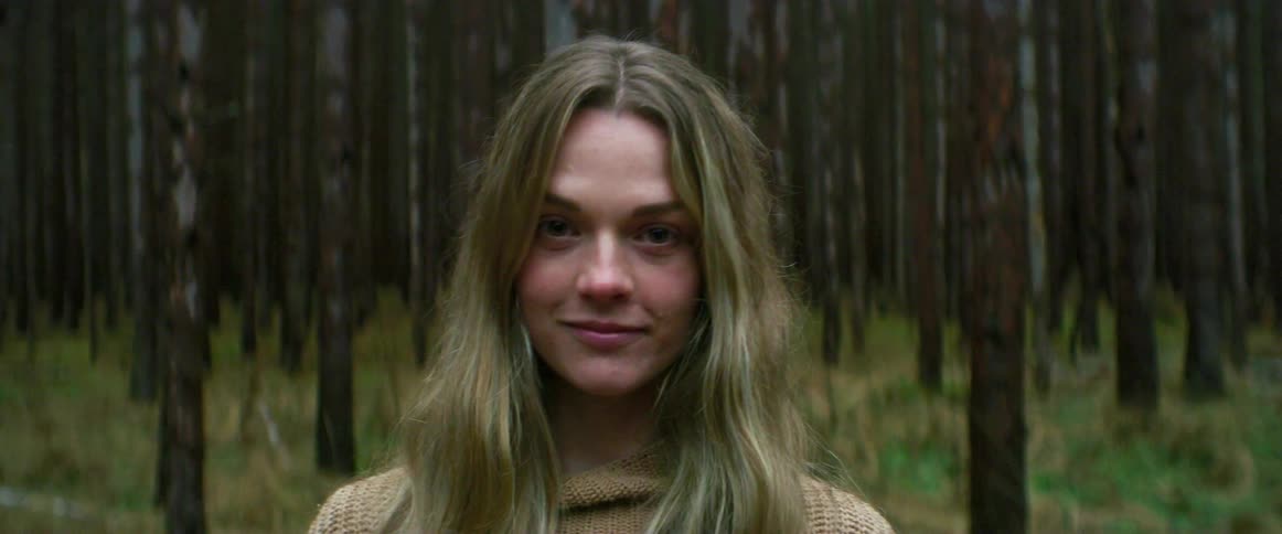 A woman standing and staring in a forest with trees and grass and removing a mask