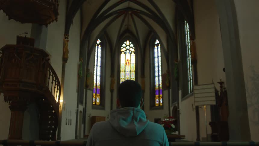 A man in a hooded sweatshirt sits alone on a pew in a church with tall stained glass windows