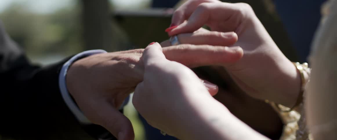 A bride slides a wedding ring onto her grooms finger as they stand together outdoors