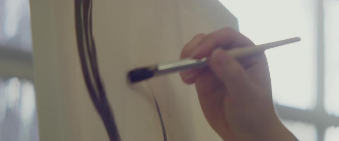 A female artist brushing dark paint onto a canvas in front of a sunny window