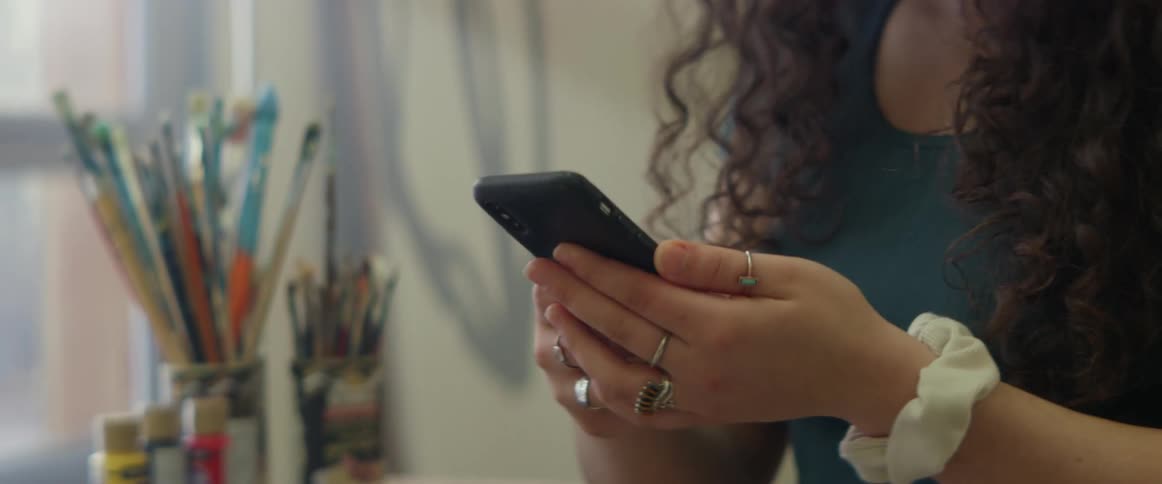 A young woman picks up her cell phone from a table that has paints and paint brushes on it