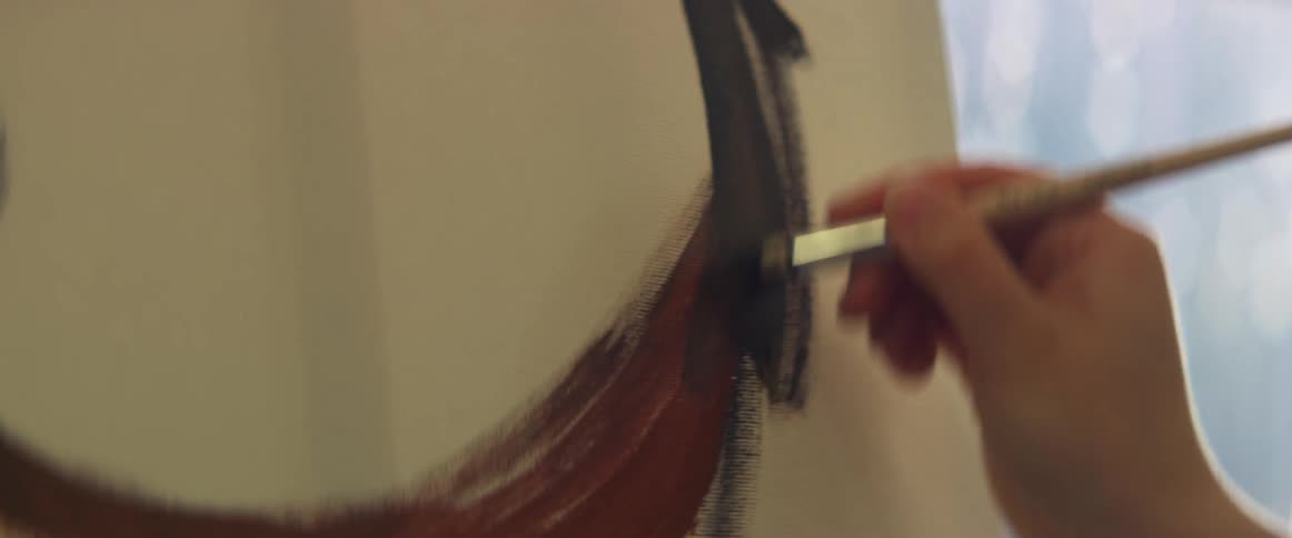 A painter uses a paint brush to make broad strokes on a canvas indoors during the day
