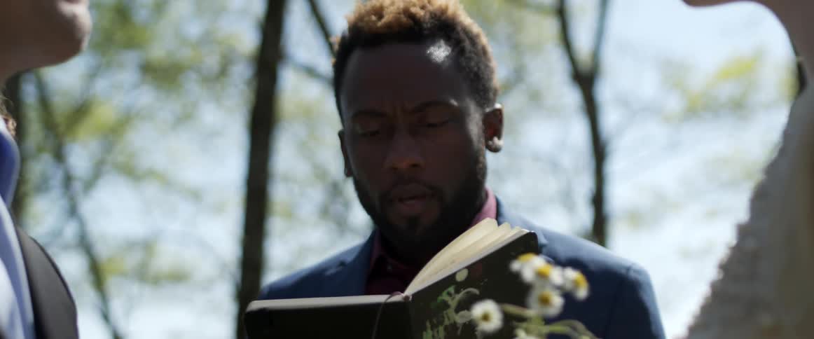A man reading from a book while conducting a wedding ceremony outside in summer