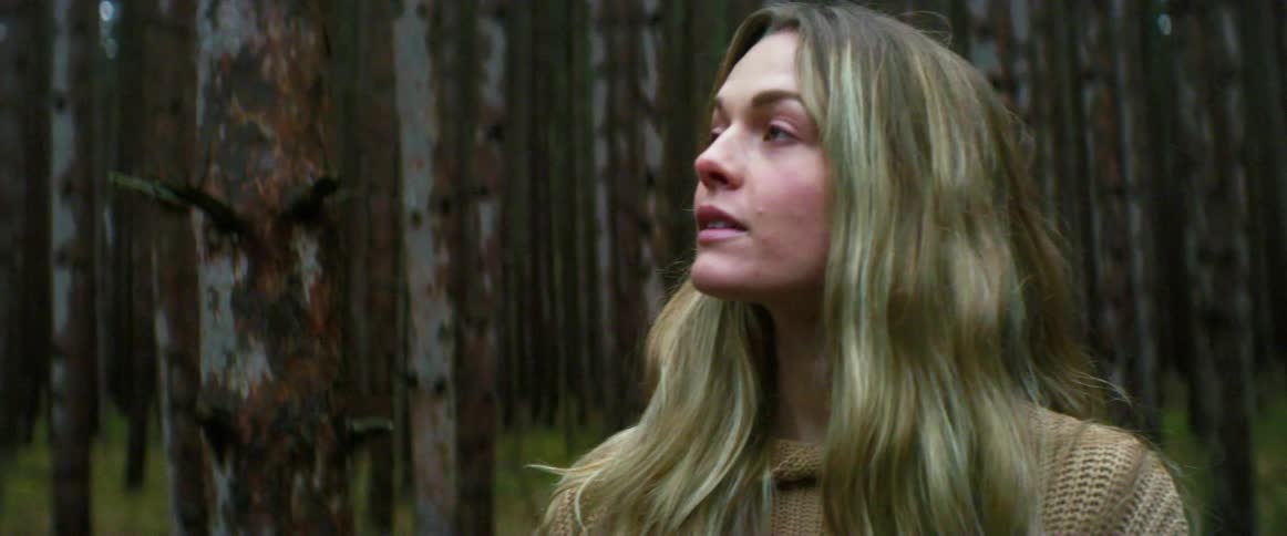 A young woman removes a medical mask from her face as she gazes up at the trees around her in a woodland during the day