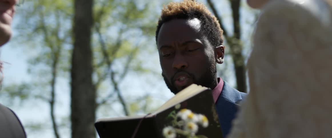 A bride and groom gaze at one another while a wedding officiant reads to them from a book as the three stand outdoors during the day