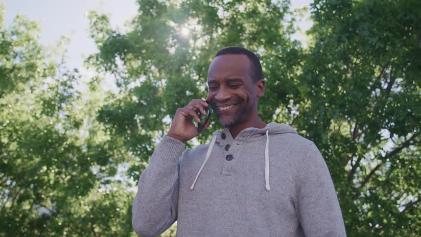 A middle aged man nods and smiles while listening to his smartphone as he stands outdoors on a sunny day