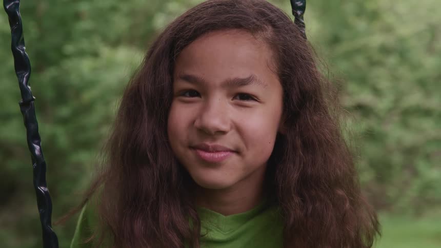 A girl sits on a swing smiling softly with trees and grass behind her