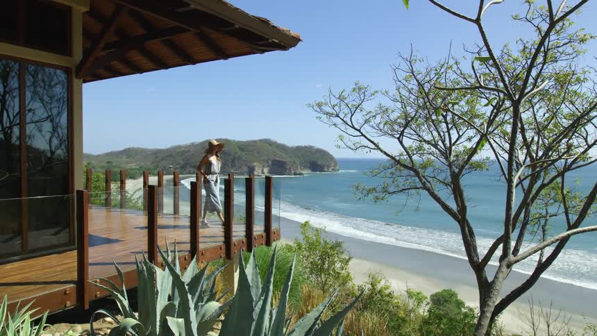 A woman looks out over the nearby beach and ocean as she walks along the balcony of a house on a sunny day