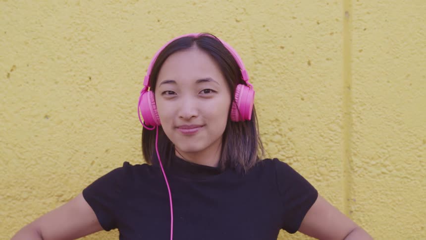 Young woman wearing pink headphones standing in front of a yellow wall looking at the camera
