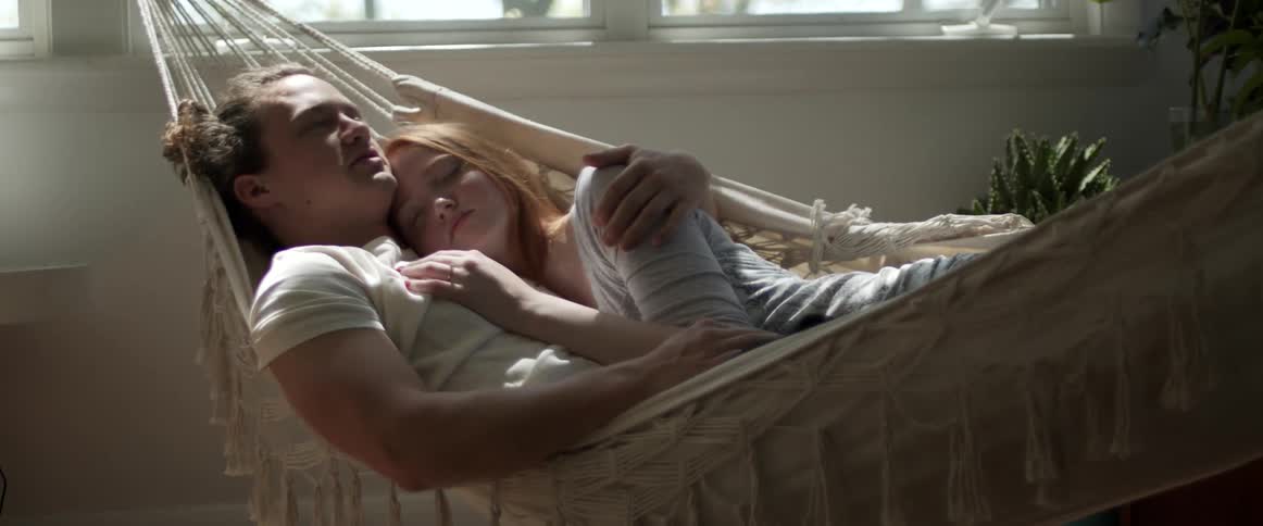 A man and a woman in a room laying and hugging on a hammock with a wall and window in the background