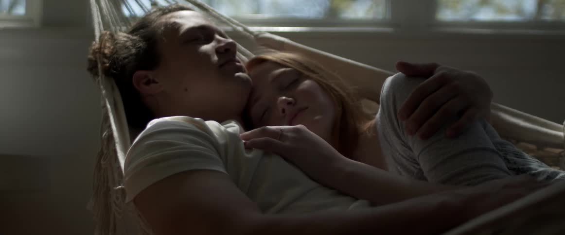 A man and a woman laying and hugging on a hammock in a room with a wall and window in the background