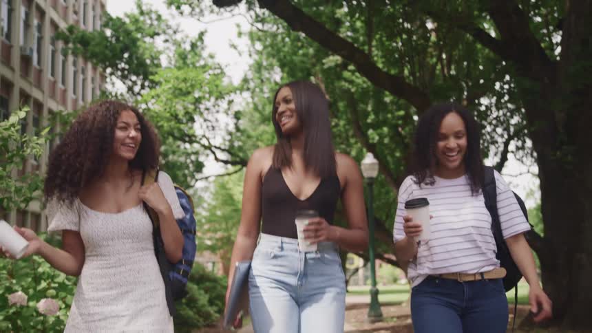 Three young women walk along a sidewalk together holding coffee cups