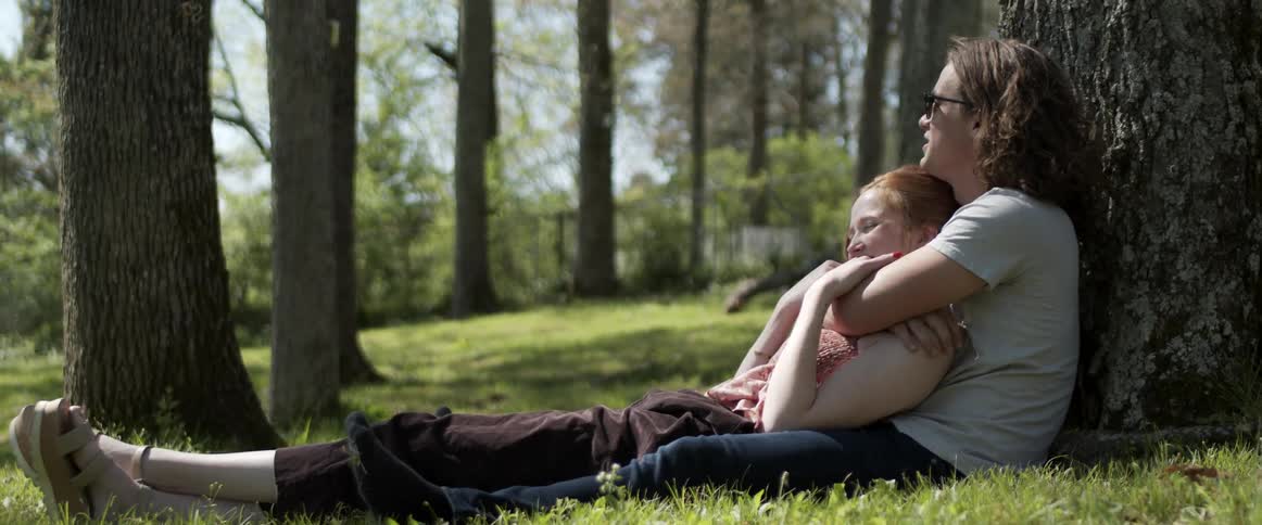 A man with his arms wrapped around his girlfriend while sitting next to a tree in summer