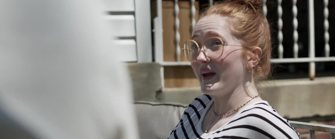 A woman with glasses and a necklace sitting and talking with a couch and a house in the background