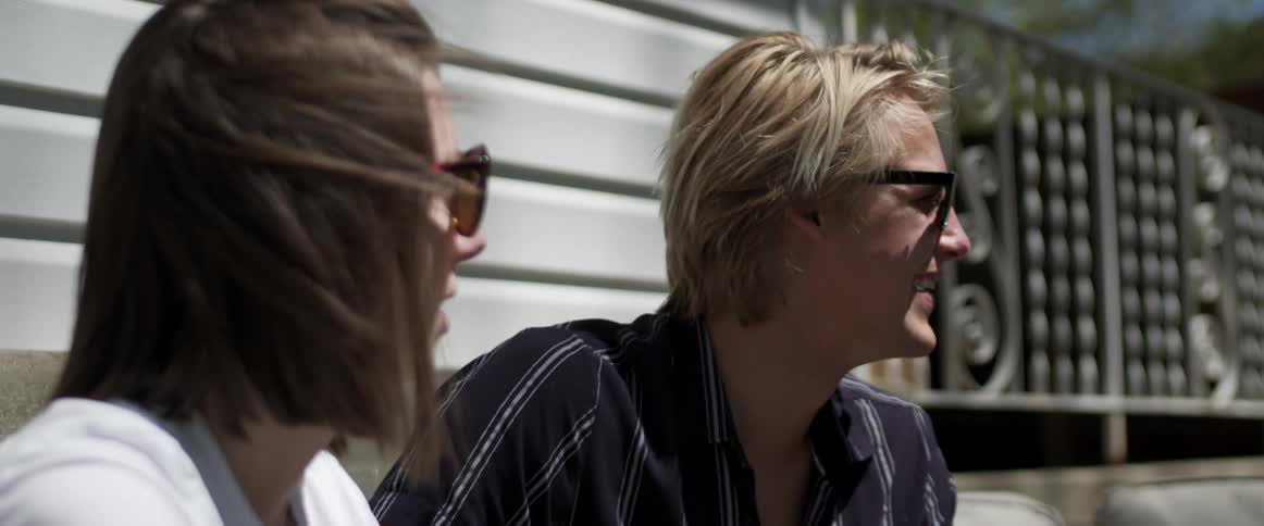 Two friends wearing sunglasses talking while sitting outside a building on a sunny day