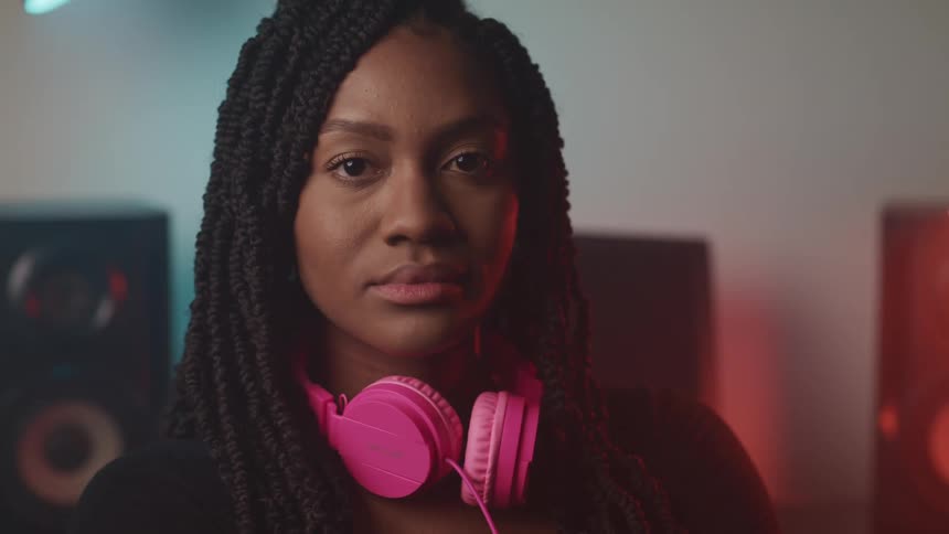 Woman with braids and pink headphones looks at the camera with speakers in the background