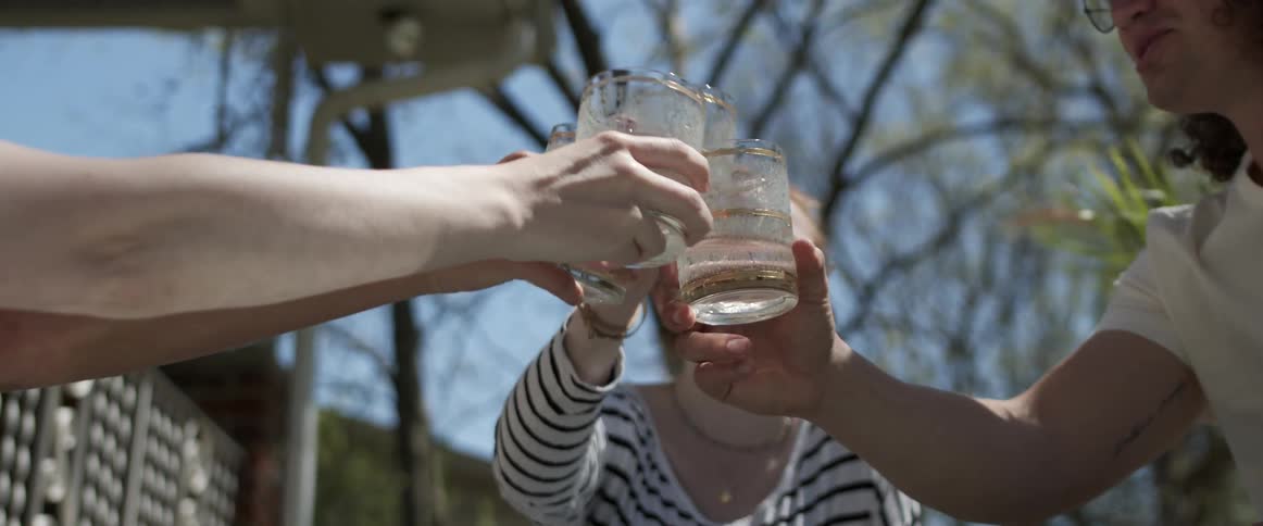 Three friends laugh as they sit outside in a semi circle and hold up their glasses to a toast