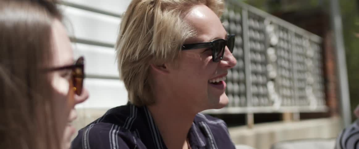 A man and two women sitting and talking with sunglasses with a house and a tree in the background