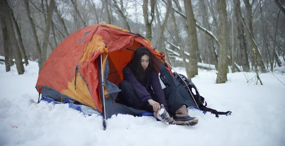 Young woman sitting on a tent in the forest at winter puts her boots on