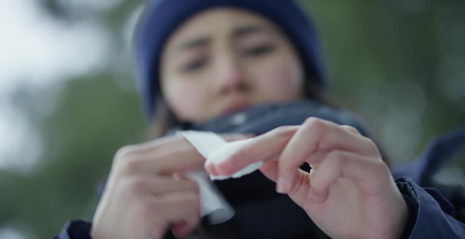 A young woman wraps a bandage around her bleeding finger while standing outdoors holding her gloves in her mouth