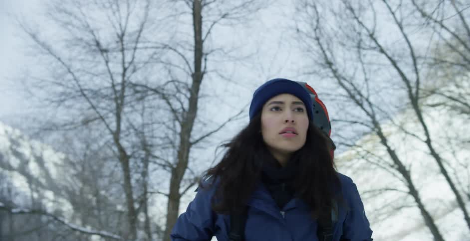 A woman with a jacket a hat and a backpack hiking with a mountain range trees the sky and clouds in the background