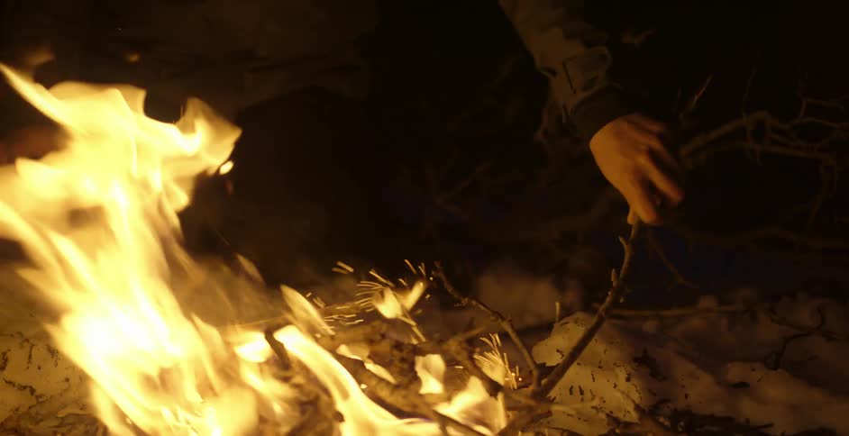A man poking a burning campfire with a stick while wearing a headlamp in the dark