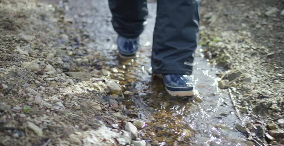 Legs in snow pants and wearing boots walk through a narrow stream of water near melting snow