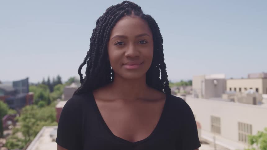A beautiful black woman smiles softly with building rooftops and trees behind her