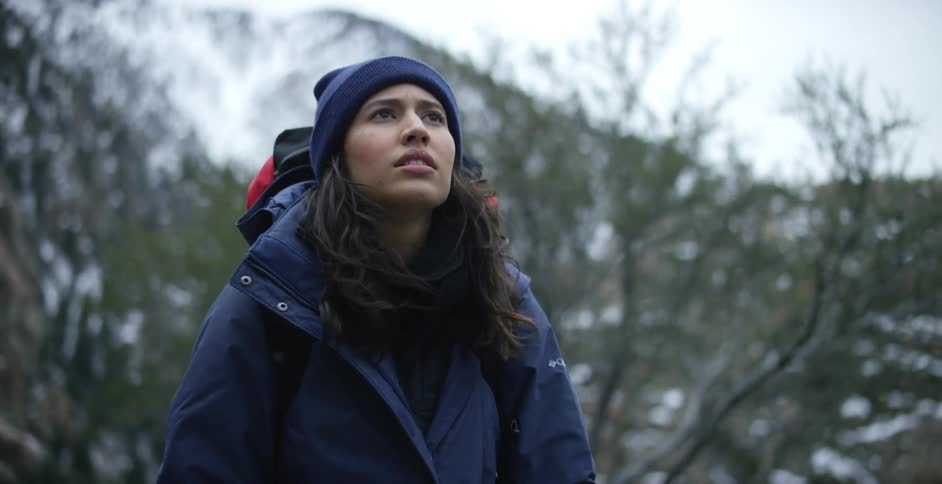 A woman with a hat coat and backpack hiking with a mountain range the sky and clouds in the background