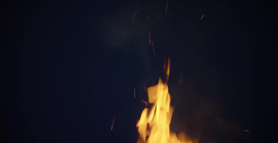The flames of a small fire flicker in the darkness of night as it lets off smoke and embers