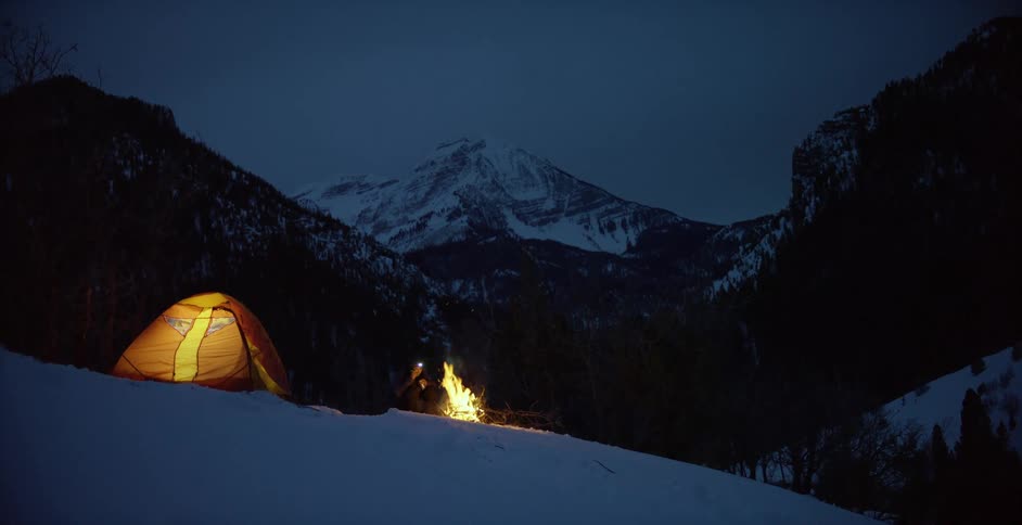 A person sitting by a campfire next to a tent with trees a mountain range the sky and clouds in the background