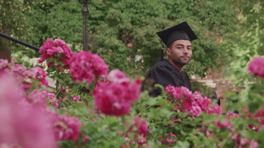 Man wearing graduation gown sits in park surrounded by flowers smiling slightly