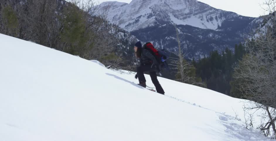A woman with a hat and backpack hiking up a snowy slope with a mountain range the sky and clouds in the background