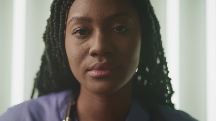 A woman with scrubs and a stethoscope in a room with windows and blinds in the background