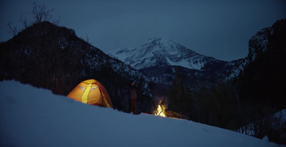 A person standing next to a campfire and a tent with trees mountains the sky and clouds in the background