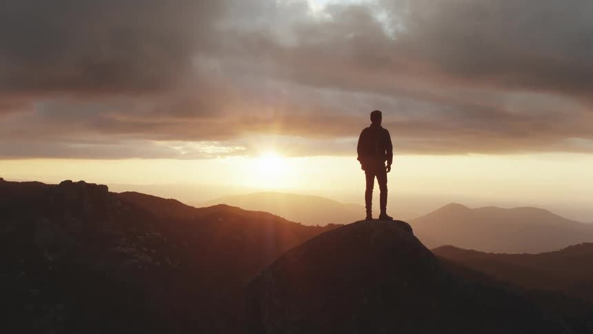 Mans silhouette stands on rock and looks around while the sun goes down