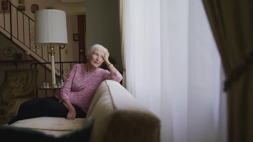 An elderly woman gazes out a window contemplatively while sitting on a sofa in the living room of a house during the day