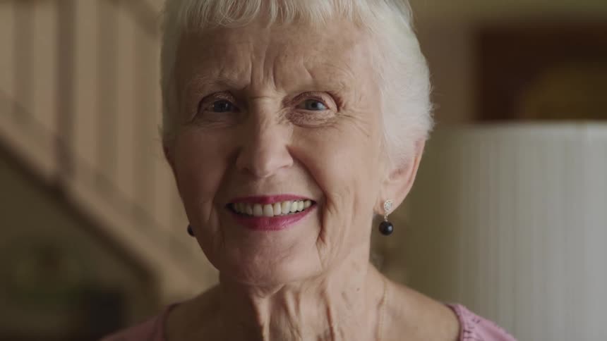 Elderly woman stands near lamp with staircase in background smiling broadly