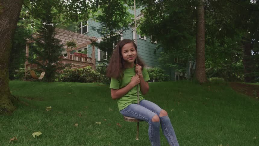 A girl sitting on a swing with trees a house the sky and clouds in the background