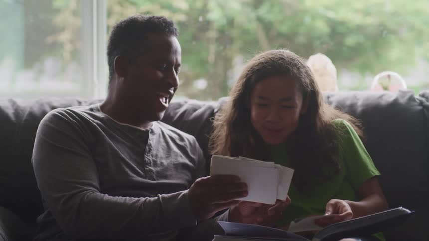 A father and daughter sit together on a couch while happily looking through photos