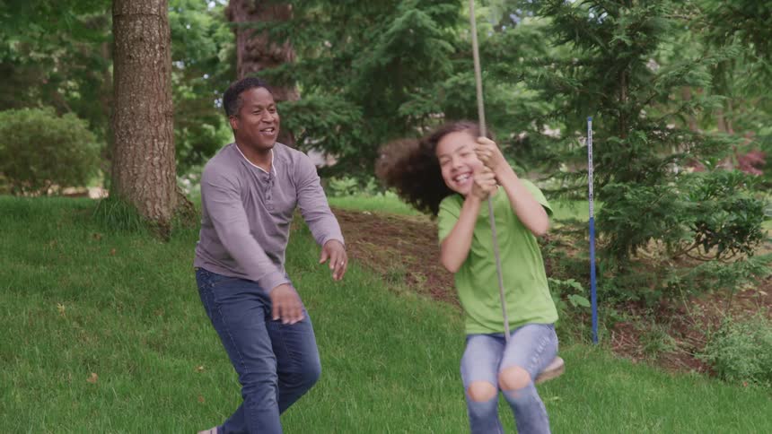 A man pushing a girl on a swing with trees the sky and clouds in the background