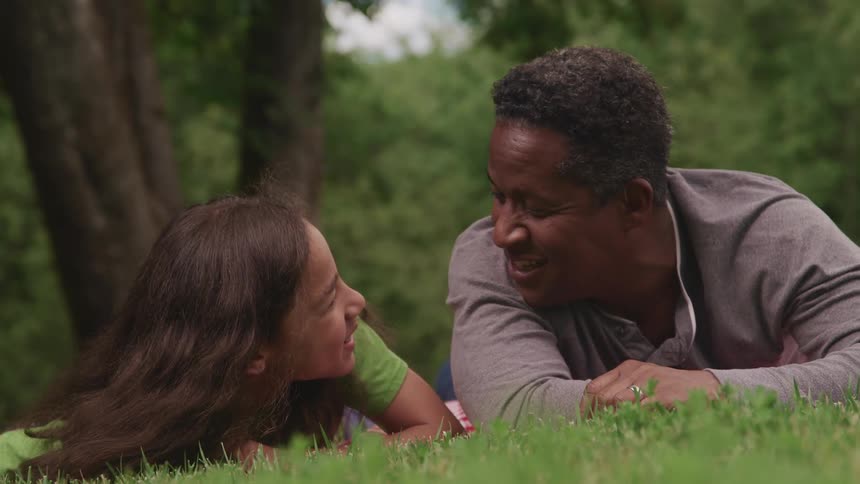 A man and a girl laying on a blanket on grass with trees the sky and clouds in the background