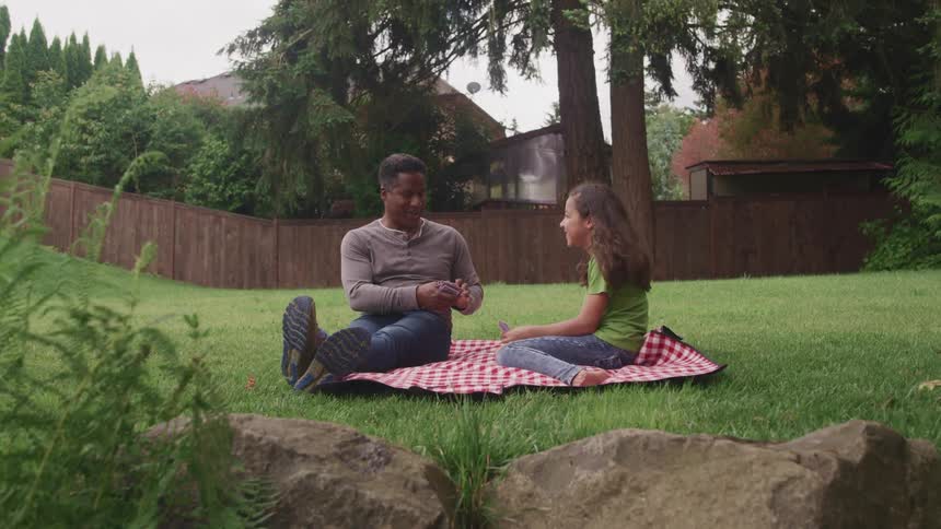 A man and a girl sitting on a blanket on grass playing cards with houses trees the sky and clouds in the background