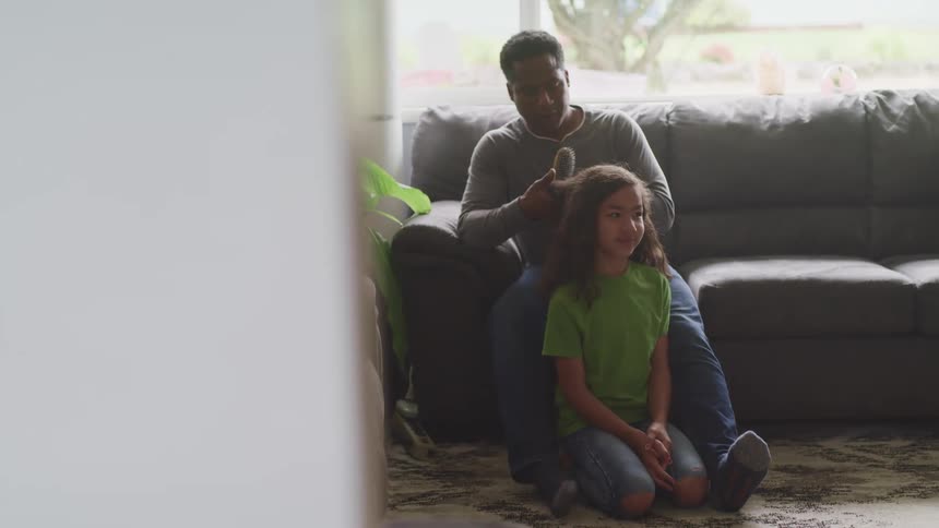Daughter kneels on floor in living room as father brushes her hair